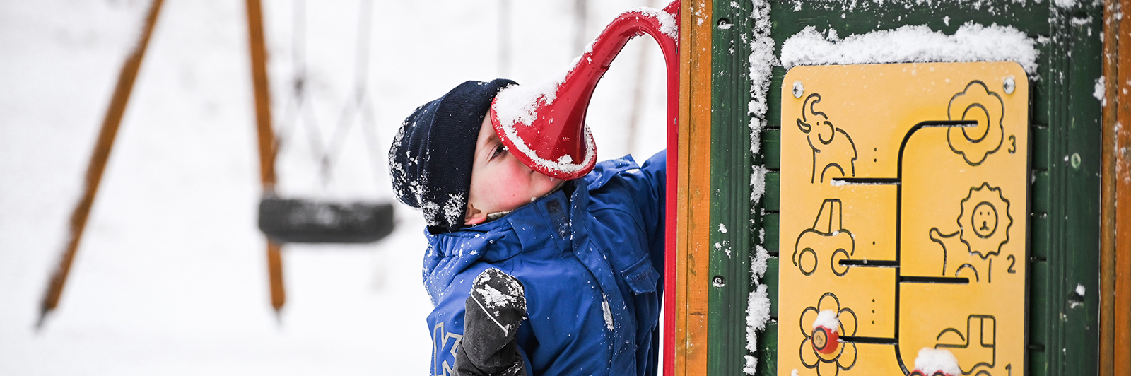 a young boy in a hat and gloves, stands in a snowy playground speaking into a talk tube
